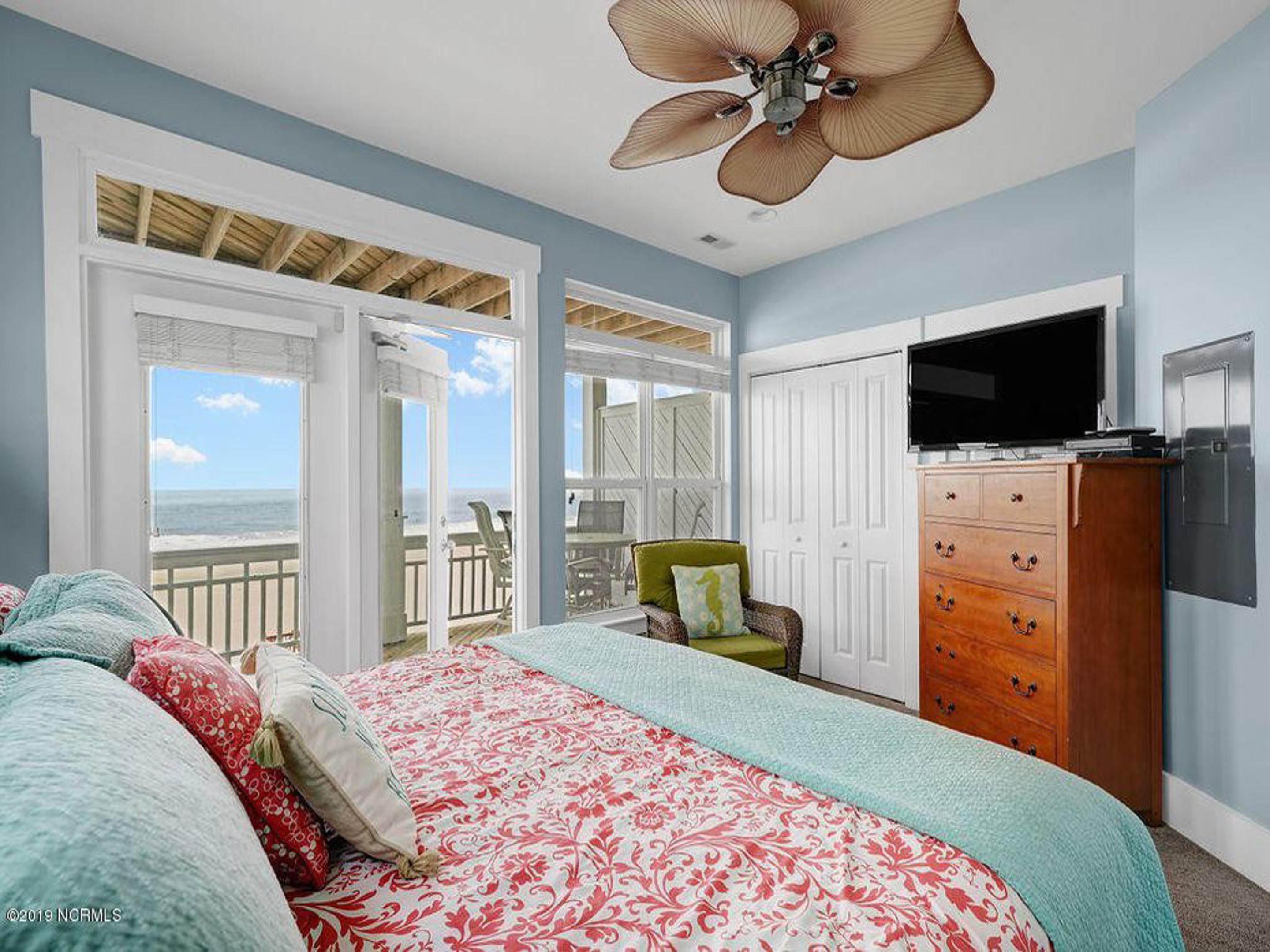 A bedroom with a bed, a dresser, a TV, and a balcony with a view of the ocean.