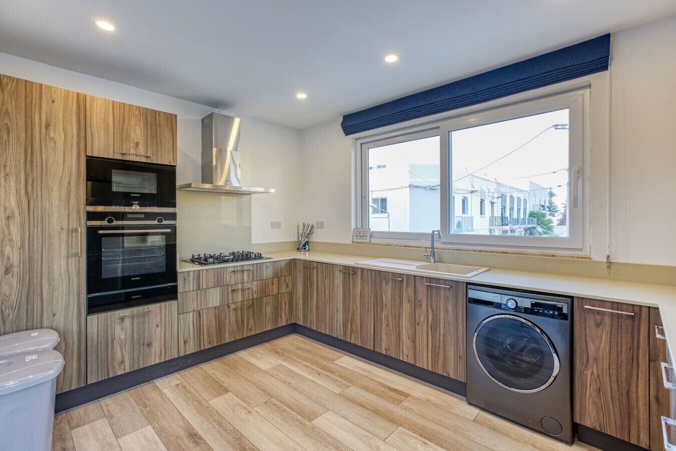 Une cuisine moderne avec des meubles en bois, un four encastré, un lave-vaisselle et une fenêtre donnant sur des immeubles.