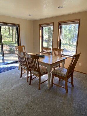 A dining area with a wooden table and chairs, large windows with a view of trees, and a carpeted floor.