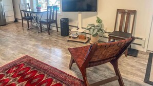 A living room with a TV, a dining table, chairs, a red patterned rug, a potted plant, and a wooden chair.