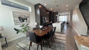A modern kitchen with dark cabinetry, a dining area with a wooden table and chairs, and a staircase leading to an upper level.
