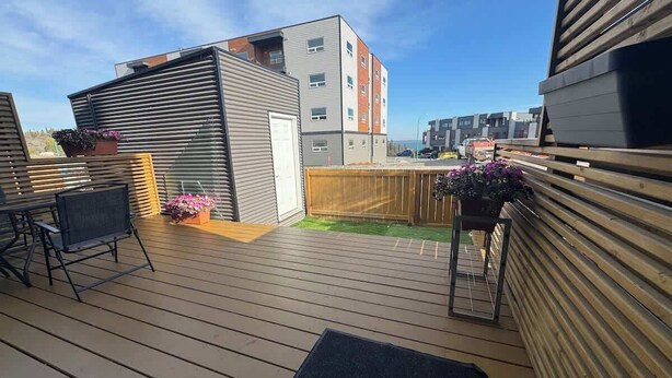 Una terraza de madera con una mesa y sillas de metal, un pequeño jardín con flores y una vista de un edificio de varios pisos al fondo.