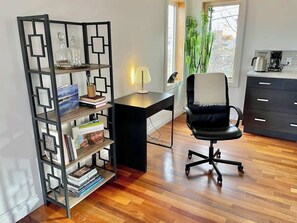 Ein Zimmer mit Schreibtisch, Bürostuhl, Bücherregal und einem Fenster mit Pflanzen.
