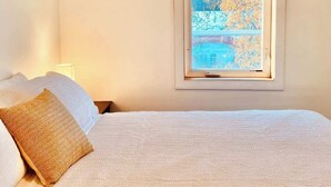 Une chambre avec un grand lit, une fenêtre donnant sur des arbres et un immeuble, et une table de chevet avec une lampe.