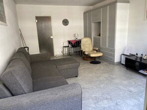 Un salon comprenant un canapé d’angle gris, un fauteuil pivotant, un bureau avec une lampe et une armoire.