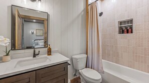 A bathroom with a sink, mirror, toilet, shower, and a shelf with toiletries. - Charming Cabin • Hot Tub & Picture-Perfect Views (Pigeon Forge)