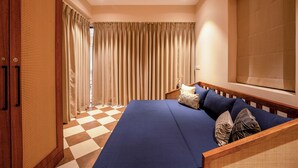 A room with a checkered floor, a bed with blue bedding, a wooden wardrobe, and a large window with curtains. - Staymaster Solintra 2BHK C101 (Verem)