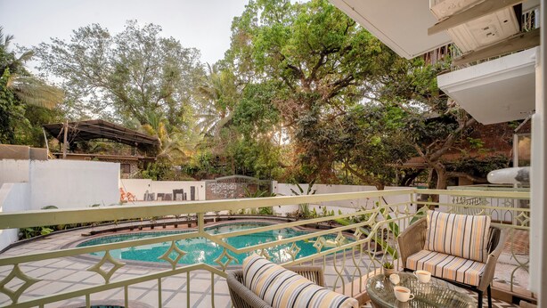 A pool area with wicker furniture and a glass table, surrounded by trees and a white building. - Staymaster Solintra 2BHK C101 (Verem)