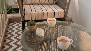 A wicker chair with striped cushions, a glass table with two white cups and a candle, and a potted plant. - Staymaster Solintra 2BHK C101 (Verem)