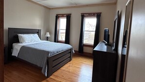Ein Schlafzimmer mit einem Bett, einem Schminktisch, zwei Fenstern mit Vorhängen und einer Decke mit Rastermuster.