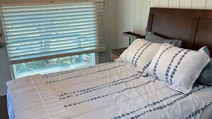 Een slaapkamer met een houten hoofdeinde, een bed met wit beddengoed en blauwe patronen, een nachtkastje en een raam met jaloezieën.