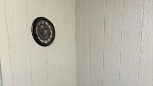 Een witte koelkast met een zwarte magnetron erop, een stopcontact aan de muur en een klok boven de magnetron.