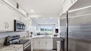 A modern kitchen with white cabinets, stainless steel appliances, a granite countertop, and a view of a green landscape through the window.