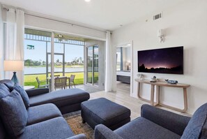A modern living room with a large flat-screen TV, grey sofas, a coffee table, and a view of a grassy area through glass doors.