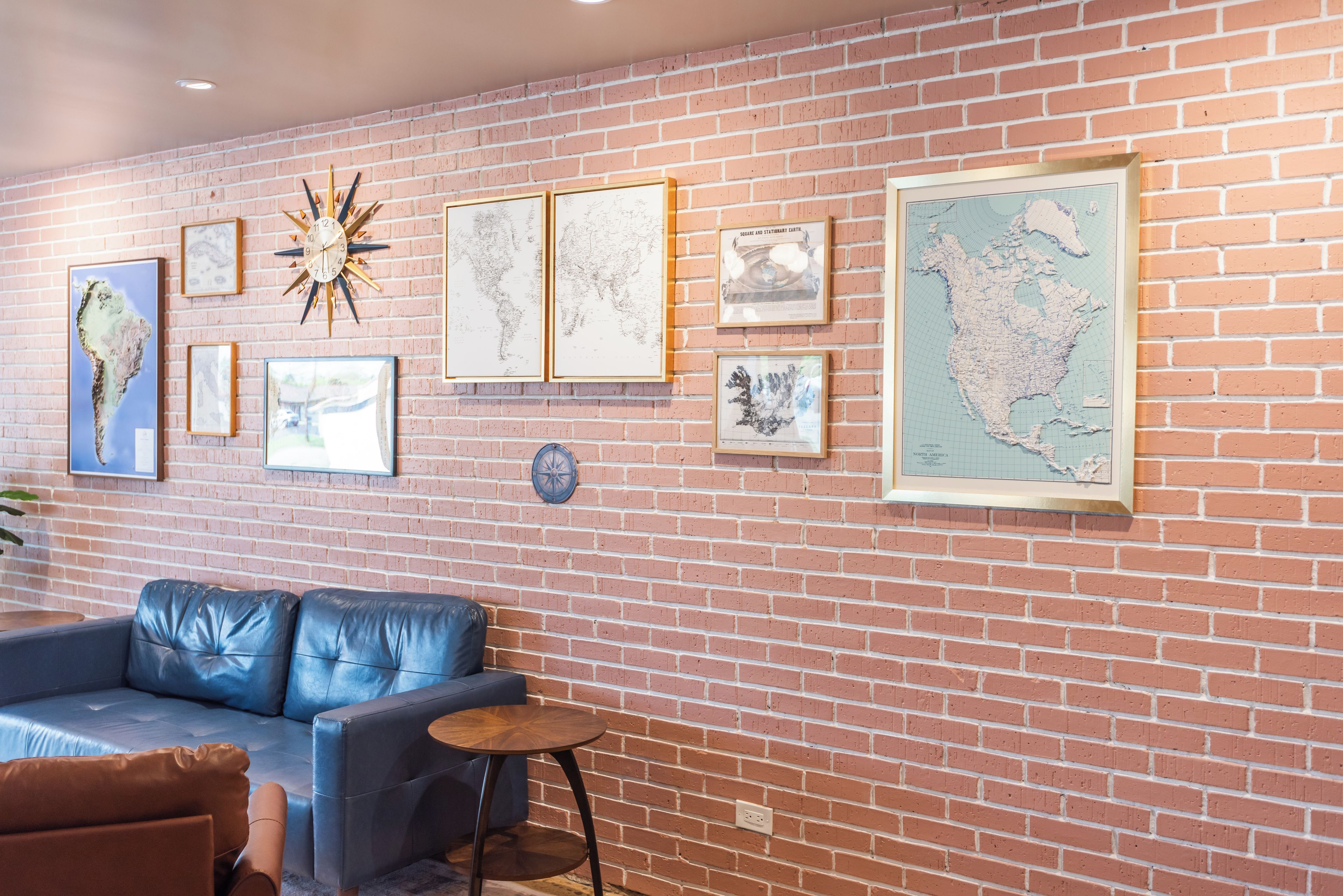 Una habitación con pared de ladrillo, un sofá de cuero azul, una pequeña mesa redonda y varios mapas enmarcados y obras de arte.