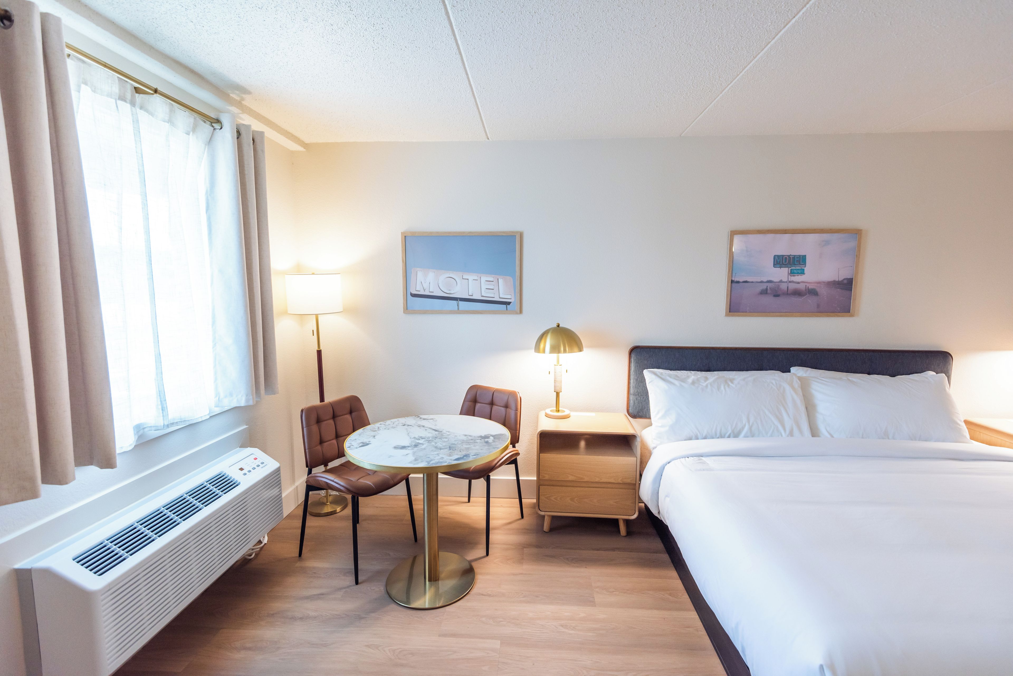 Una habitación de hotel con una cama, una mesa redonda, dos sillas, una mesita de noche, una lámpara, un aire acondicionado de pared y dos cuadros enmarcados.