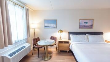 Una habitación de hotel con una cama, una mesa redonda, dos sillas, una mesita de noche, una lámpara, un aire acondicionado de pared y dos cuadros enmarcados.