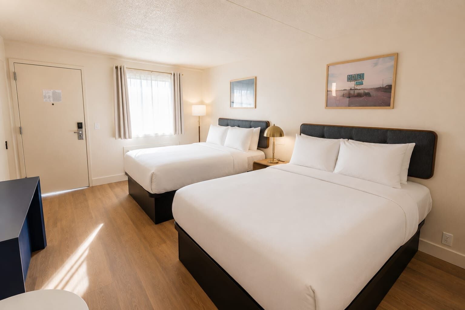 A hotel room with two beds, a desk, a lamp, and a painting on the wall.
