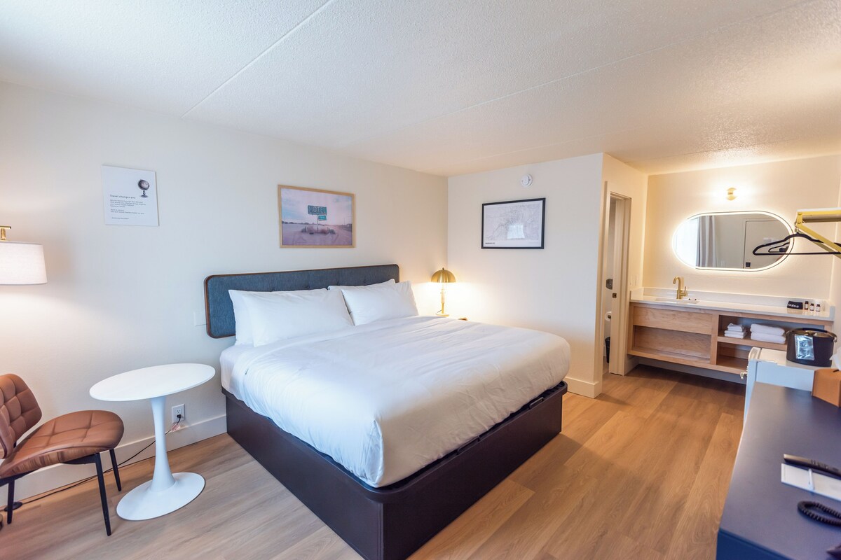 A hotel room with a bed, a bedside table, a chair, a small round table, a mirror, a sink, and a makeup station.