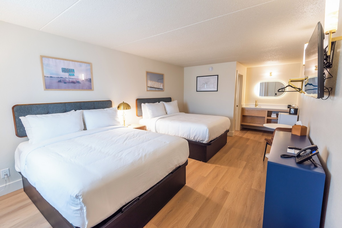 A hotel room with two beds, a desk with a telephone, a mirror, and a wall-mounted TV.