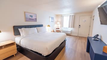 A hotel room with a bed, a desk, a chair, a lamp, a window with curtains, and a door.