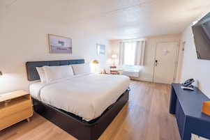 A hotel room with a bed, a desk, a chair, a lamp, a window with curtains, and a door.