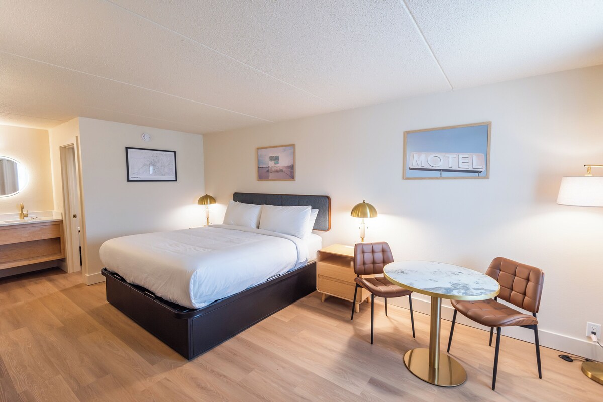 A hotel room with a bed, bedside table, lamp, chair, and a small round table.