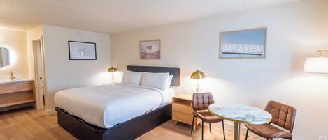 Una habitación de hotel con cama, mesita de noche, lámpara, silla y una pequeña mesa redonda.
