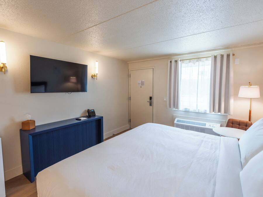 Habitación de hotel con cama, escritorio con teléfono, televisión de pantalla plana en la pared y ventana con cortinas.