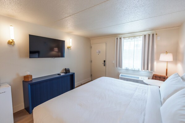 A hotel room with a bed, a desk with a telephone, a flat-screen TV mounted on the wall, and a window with curtains.