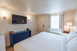 Habitación de hotel con cama, escritorio con teléfono, televisión de pantalla plana en la pared y ventana con cortinas.