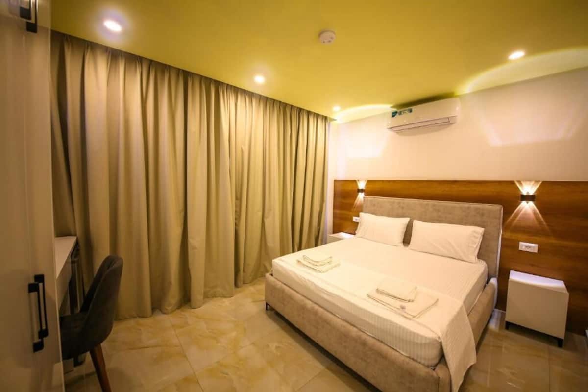 Une chambre d’hôtel équipée d’un lit, de rideaux, d’un bureau, d’une chaise, d’une table de chevet, d’un climatiseur et d’une lampe fixée au mur.