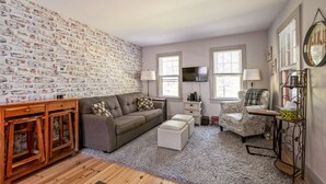 Ein Wohnzimmer mit Backsteinwand, einem grauen Sofa, einem gemusterten Sessel, einem Holzschrank und einem Fernseher an der Wand.