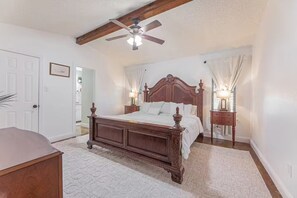 Une chambre avec un lit en bois, un ventilateur de plafond, deux lampes de chevet et une porte sur le côté gauche.