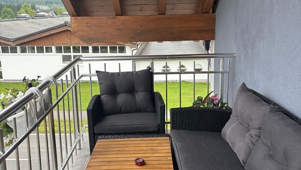 A covered balcony with a wooden ceiling, two black sofas, a wooden table, and a metal railing. - SC Apartments (Bad Goisern am Hallstättersee)