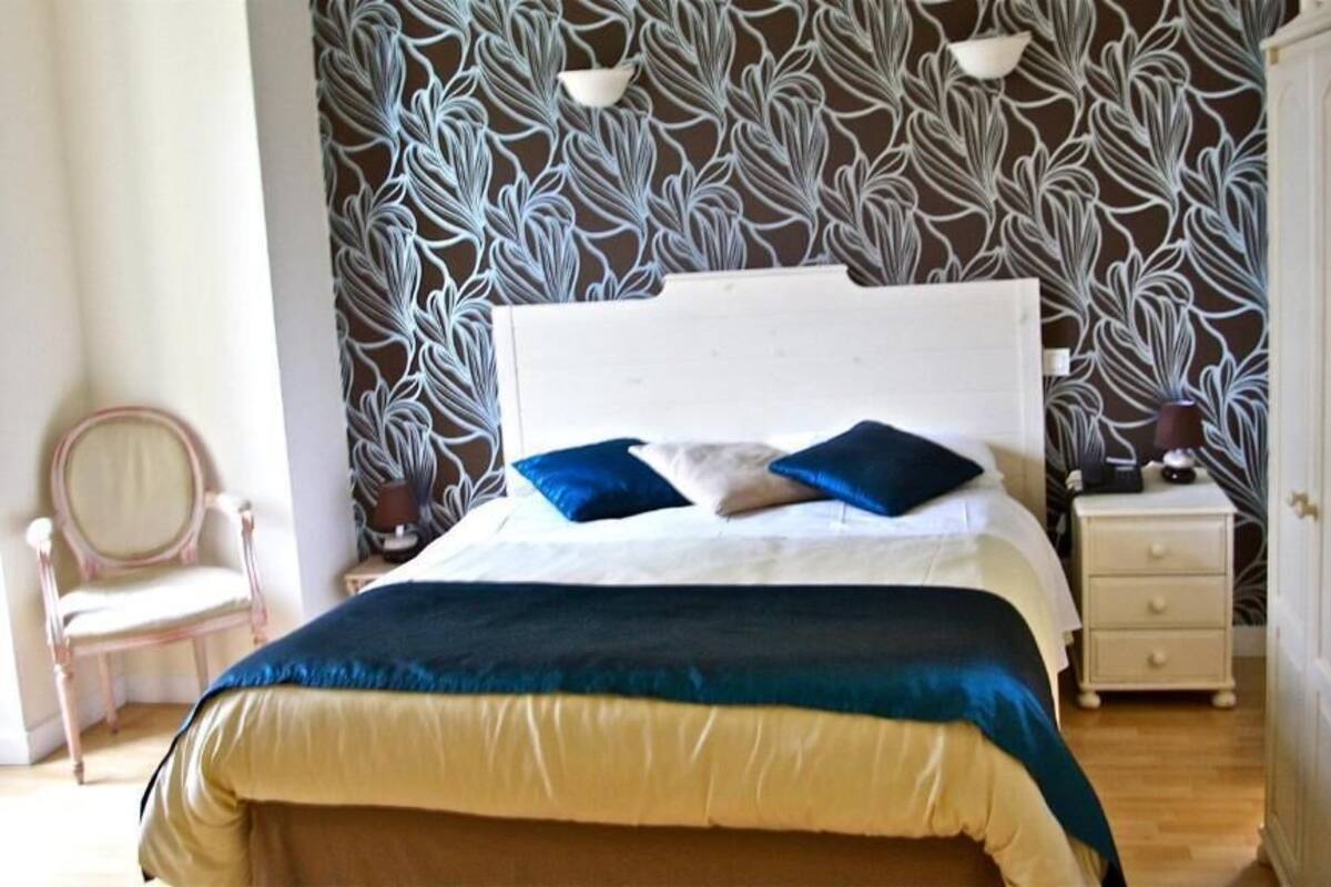 A bedroom with a bed, bedside table, lamp, chair, and a patterned wallpaper.