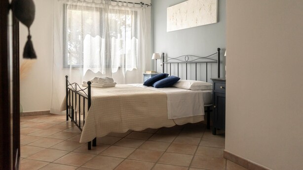 A bedroom with a bed, a nightstand, a lamp, a window with curtains, and a wall-mounted artwork. - Country House Frantoiano (ROSIGNANO MARITTIMO)