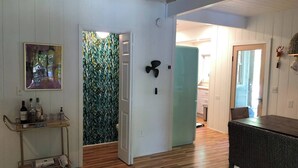 Ein Zimmer mit Holzboden, einer Glastür zu einem weiteren Raum, einem Wandventilator und einem kleinen Tisch mit Stuhl.