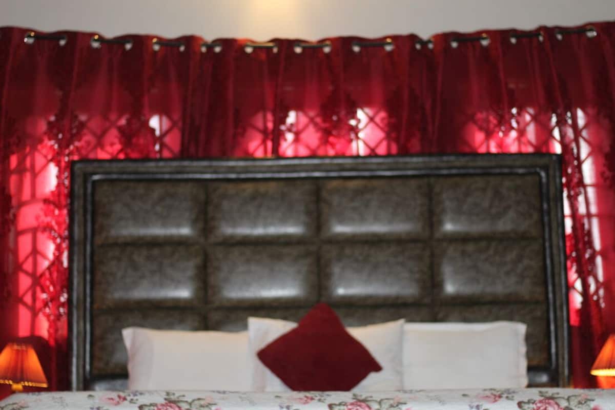 A bedroom with a large headboard, a floral bedspread, and red curtains.
