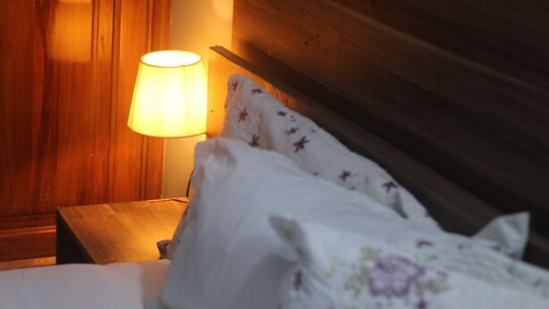 Cama com roupa de cama branca e padrão floral, mesa de cabeceira de madeira com abajur e cabeceira de madeira.