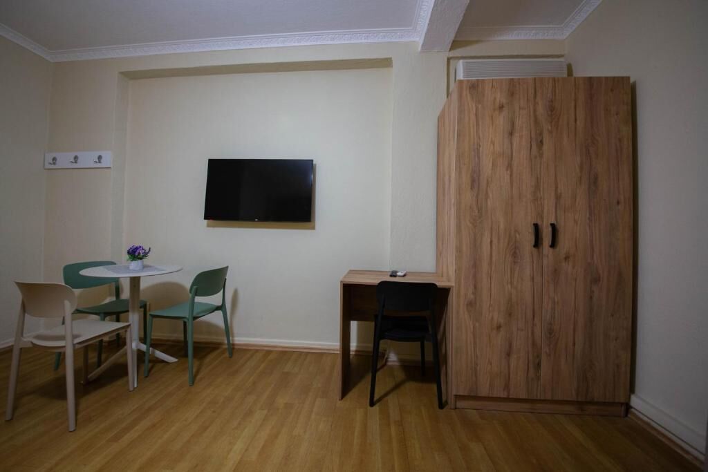A room with a wooden wardrobe, a TV mounted on the wall, a small round table with chairs, and a wooden desk with a chair.