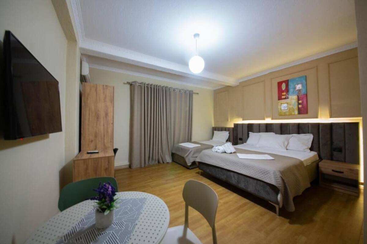 Una habitación de hotel con cama, mesitas de noche, un escritorio con silla, una mesa de comedor con un jarrón y un televisor montado en la pared.