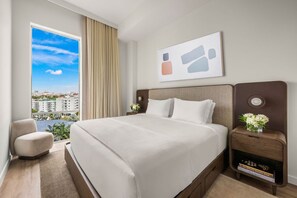 Une chambre d’hôtel avec un grand lit, une vue sur la ville, une fenêtre avec des rideaux, une table de chevet ornée d’un vase de fleurs et une œuvre d’art accrochée au mur.