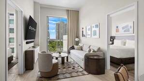 Et moderne hotelværelse med sofa, lænestol, sofabord og seng. Der er et stort vindue med udsigt over byen.