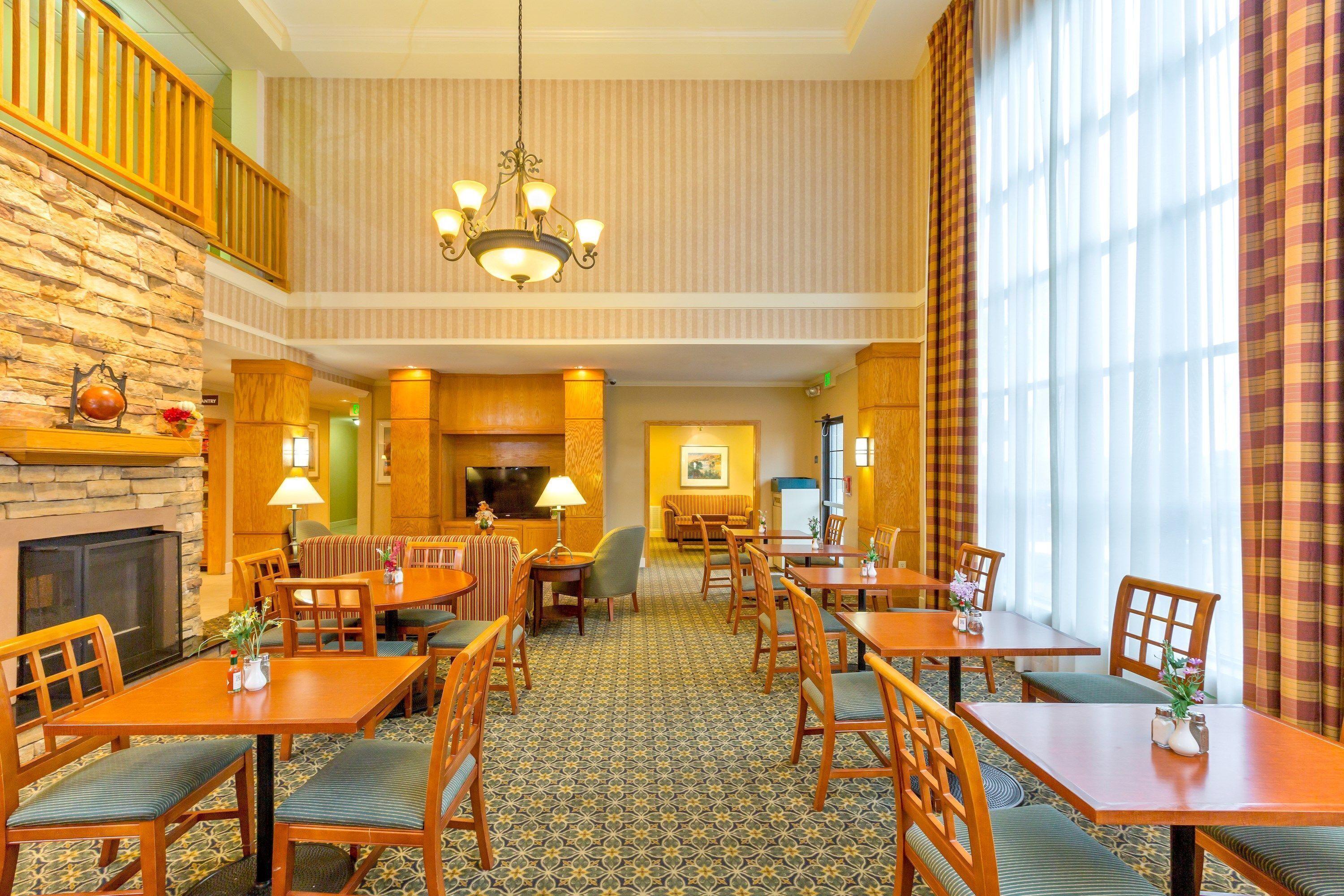 A hotel lobby with multiple dining tables and chairs, a fireplace, a chandelier, and a large window with curtains.