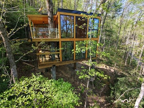 Ein Baumhaus mit einer Holzterrasse und großen Fenstern, umgeben von Wald.