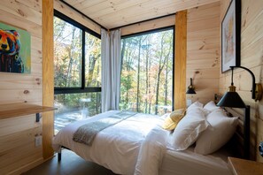 Ein Schlafzimmer mit großem Fenster, Holzwänden, einem Bett mit weißer Bettwäsche, einer Nachttischlampe und einem Gemälde an der Wand.