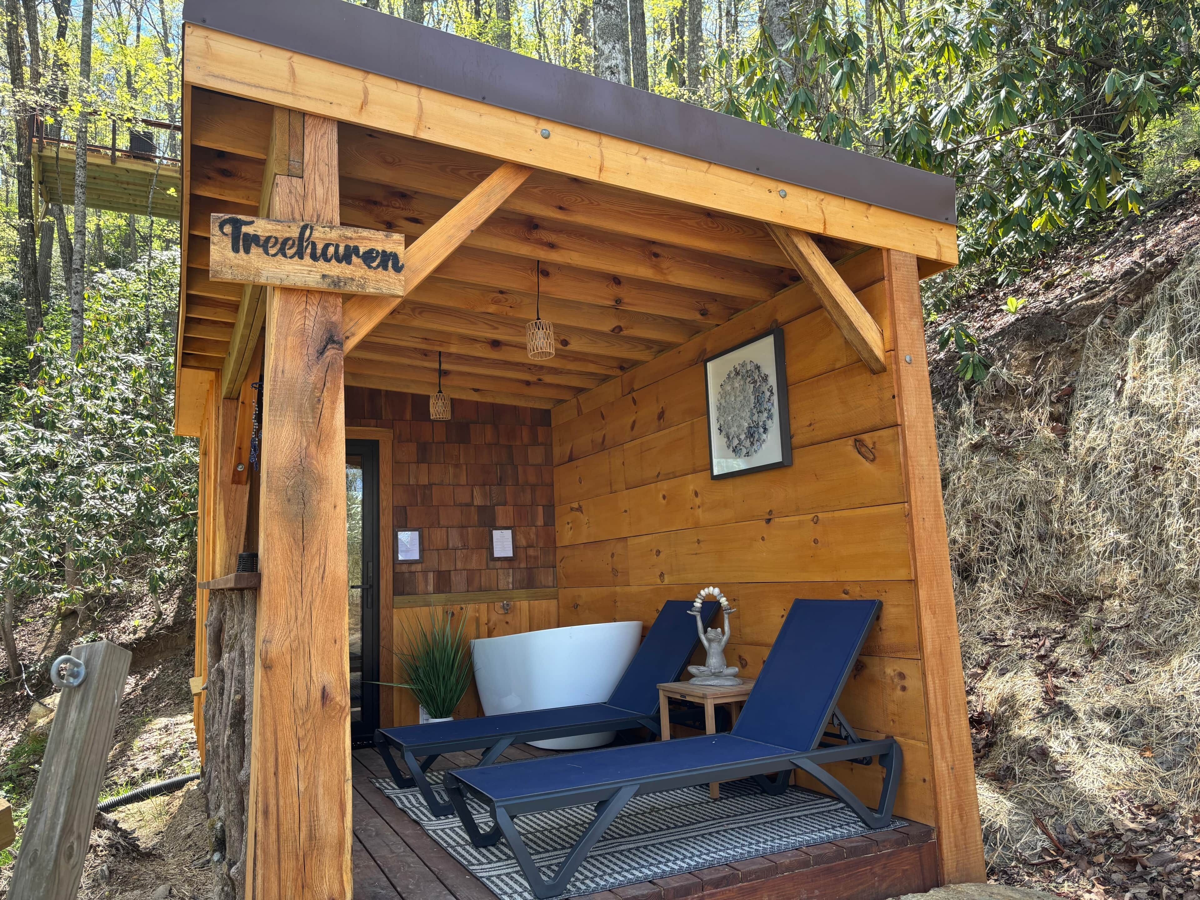 A wooden cabin labeled 'Treehaven' with a small porch, two lounge chairs, a table, and a framed picture on the wall.
