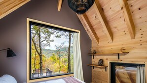 Quarto com uma janela grande, vigas de madeira no teto, cama, mesa de cabeceira e lareira.