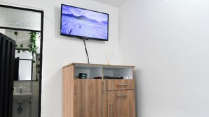 Une chambre à coucher comprenant un lit, une armoire en bois, un téléviseur fixé au mur, et une salle de bain avec un lavabo et un miroir.